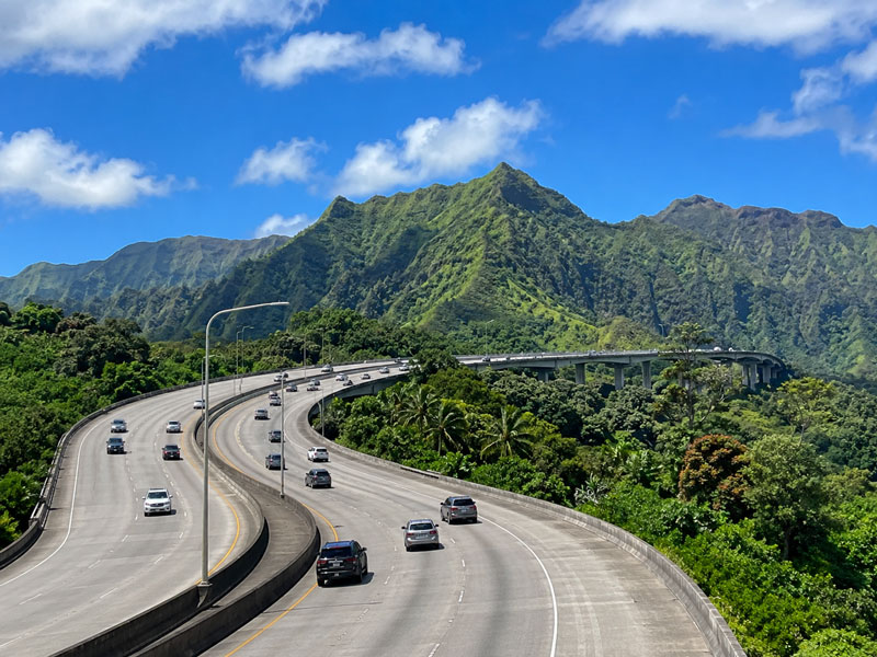An elevated winding multi-lane highway viaduct curving through lush tropical mountain valleys under a vibrant blue sky.