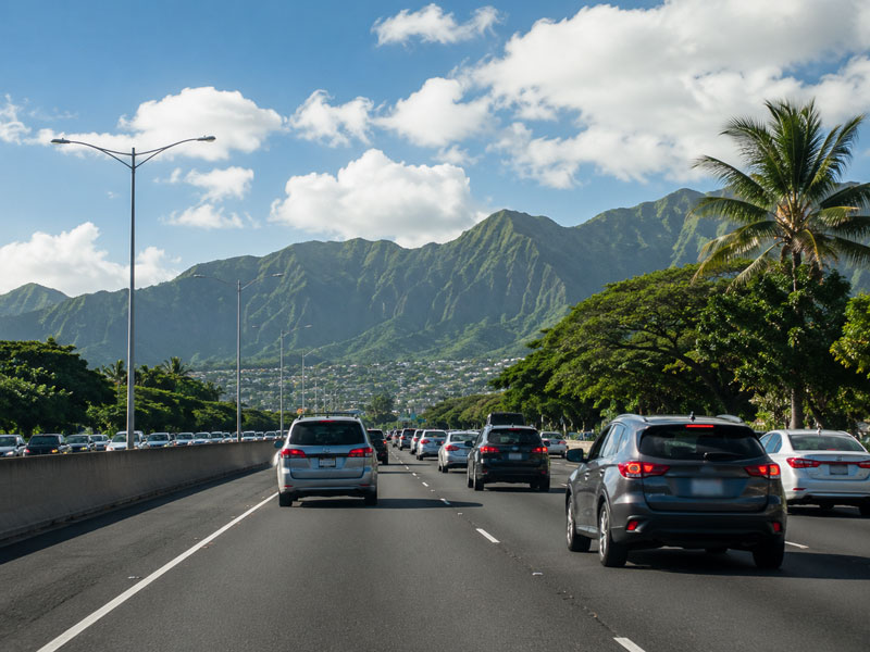 Cars driving on a multi-lane highway in Hawaii with lush green mountains and palm trees under a sunny sky.