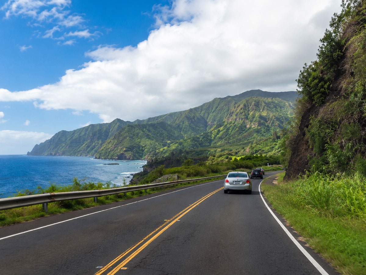 Cars driving along a cliffside coastal highway winding past dramatic volcanic cliffs, green mountains, and the deep blue Pacific ocean.