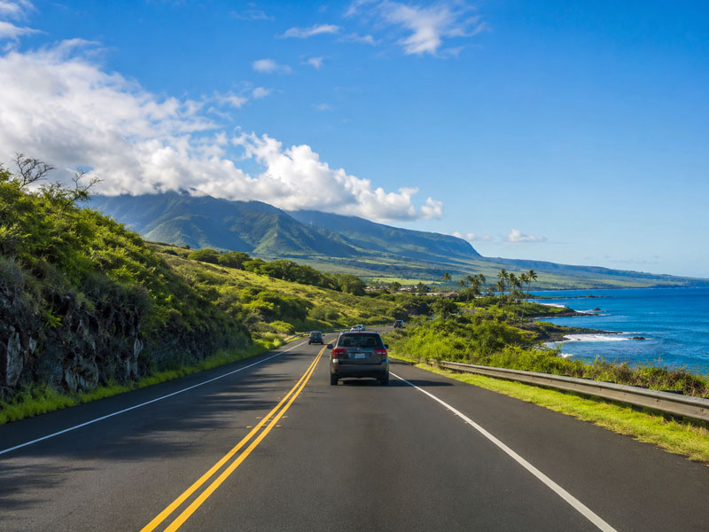 An SUV driving along a two-lane coastal highway bordered by green hillsides on one side and a deep blue ocean with palm trees on the other.