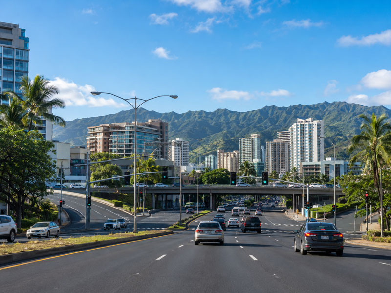 Vehicles driving on a multi-lane urban street leading toward a highway overpass, flanked by high-rise buildings and tall green mountains.