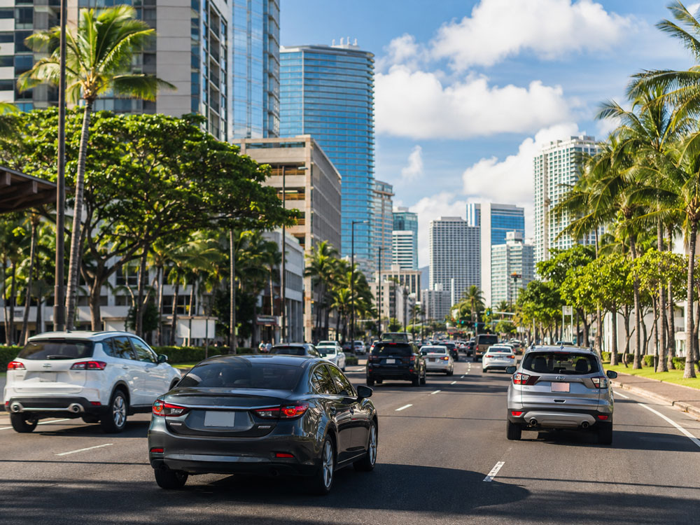 Vehicles driving along a multi-lane city street flanked by palm trees and modern high-rise skyscrapers under a blue sky.