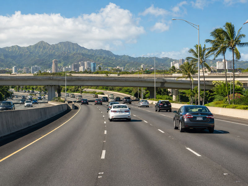 Vehicles driving on a multi-lane highway approaching an overpass bridge framed by tall palm trees, city high-rises, and green mountains.