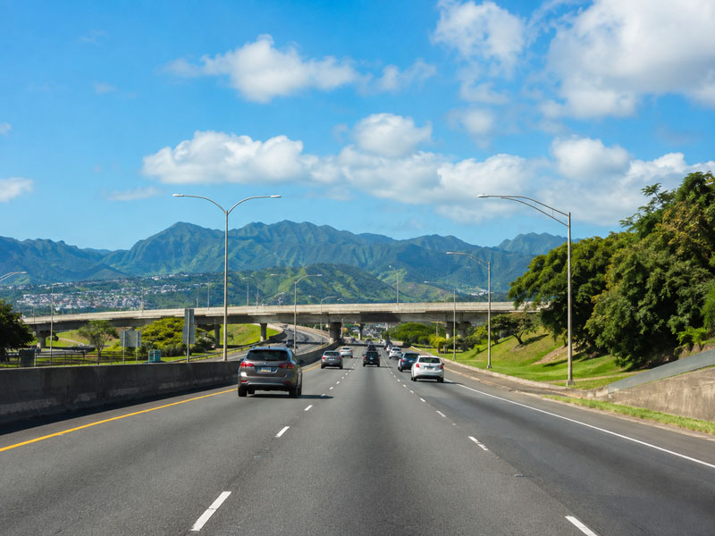 Vehicles driving along a wide multi-lane highway toward a concrete overpass with steep green mountain ridges under a blue sky with fluffy white clouds.