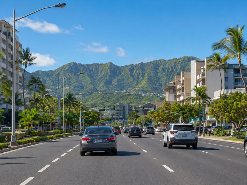 Cars driving down a multi-lane city street lined with modern apartment buildings and palm trees toward green mountain slopes under a clear blue sky.