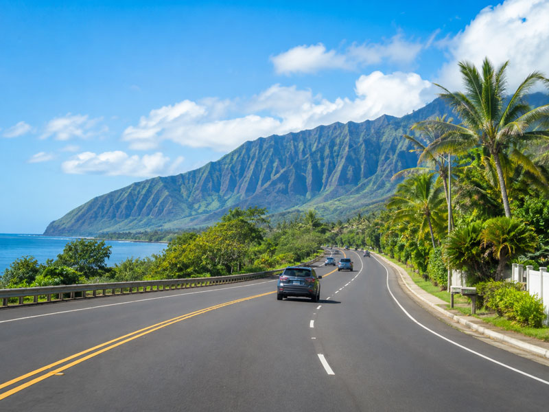 SUV driving along a paved coastal highway flanked by dramatic green mountain ridges, palm trees, and clear blue ocean in Hawaii.